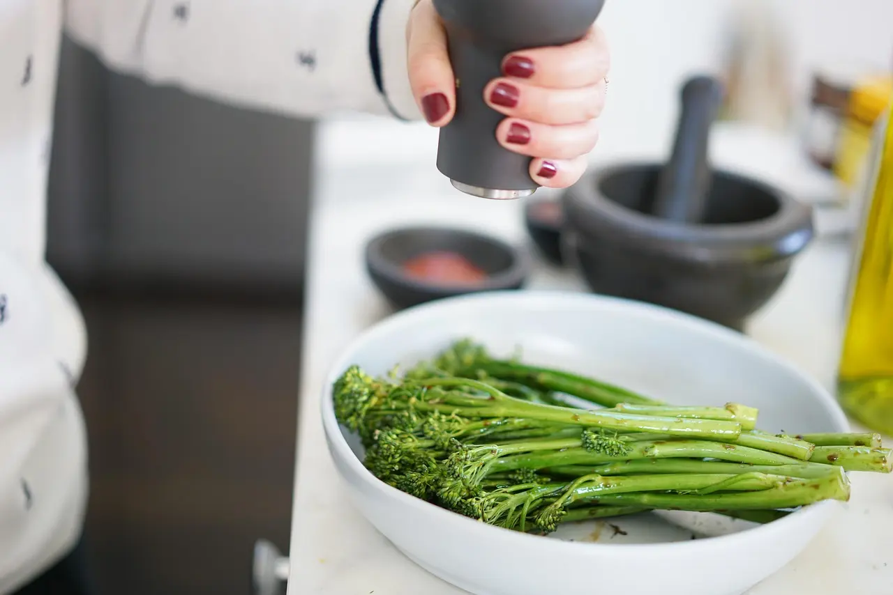 Fresh broccolini being seasoned with black pepper from a grinder, placed in a white ceramic bowl on a kitchen counter.
