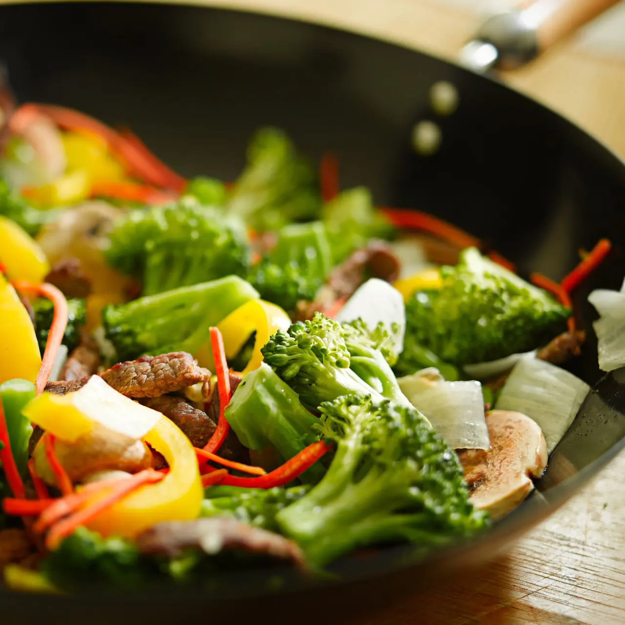 A stir-fry featuring broccolini, yellow peppers, and slices of meat cooking in a dark wok. You can see mushrooms and carrots mixed throughout, looking amazing even without sauce on a wooden table.